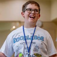 Laughing 5th-grade boy in a graphic T-shirt with a blue lanyard on, standing in a hallway of a large building